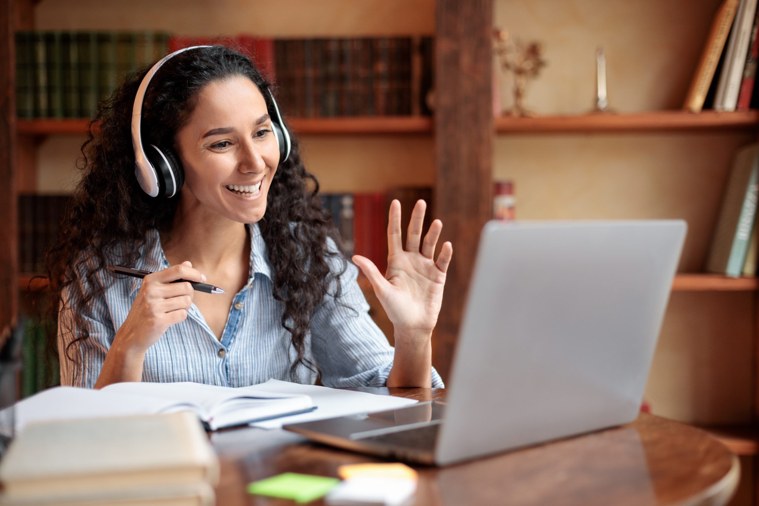 Teleconference And Telecommuting. Portrait of smiling woman sitting at desk, having video call on laptop, waving to webcam. Distance Education. Positive lady wearing headest at virtual meeting