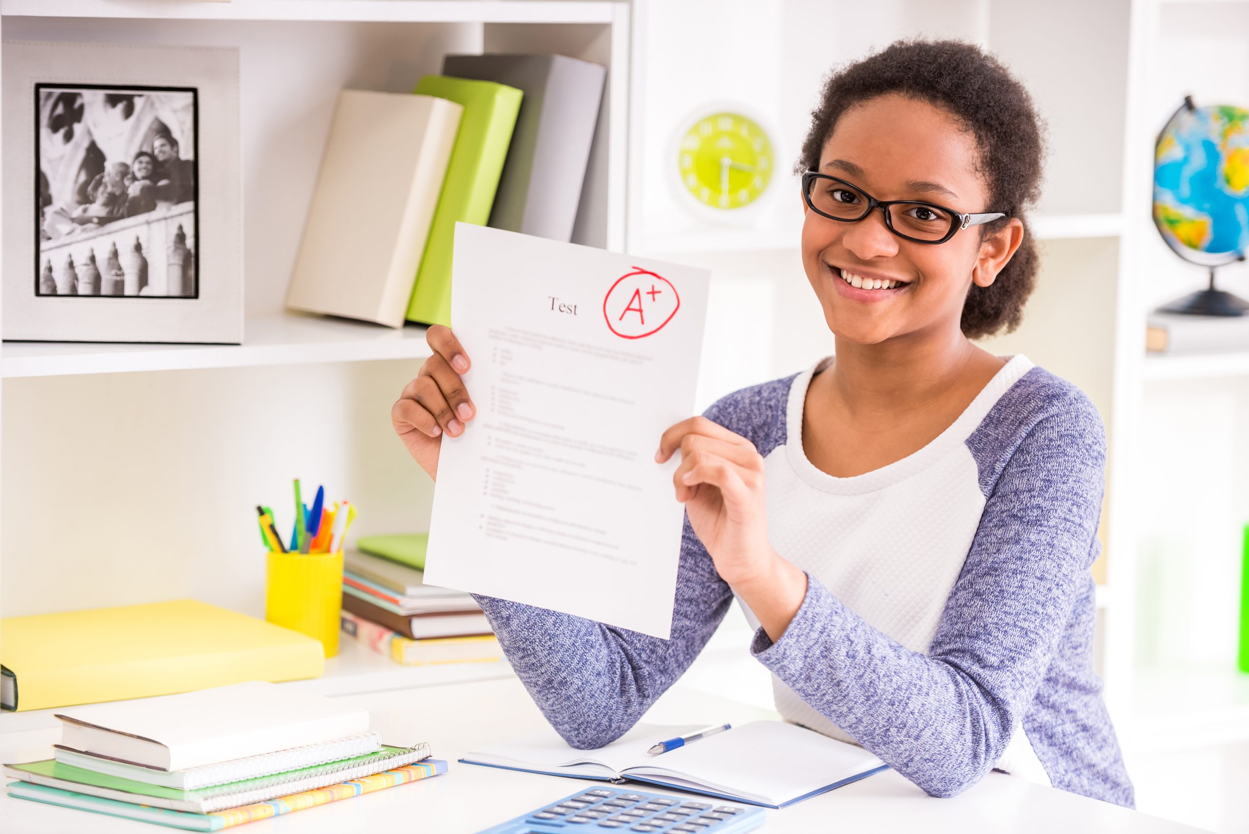 Young mulatto schoolgirl  in glasses sitting at the table and  showing perfect test results on colorful background.